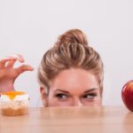 A woman peeks over a table, reaching for a small dessert beside a red apple—pondering appetite suppressant drops vs pills.