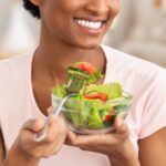 Smiling woman in a pink shirt holding a glass bowl of salad, enjoying a forkful of lettuce, cucumber, and tomato—perfect for eating a salad before bed.