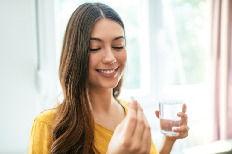 Collagen Vs. Fish Oil: Which One Is Right For You? A smiling woman in a yellow shirt holds a glass of water and a pill, contemplating the choice between collagen vs fish oil, as she prepares to take her supplement in a bright room.