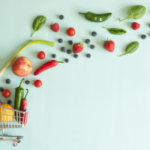 A small shopping cart holds fruits and vegetables, with IBS grocery list staples like berries, tomatoes, and greens spilling out onto a light blue background.