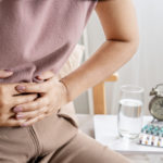 A person clutching their stomach in pain sits by a table with a glass of water, pills, and an alarm clock, wondering: what are the 3 types of IBS?.