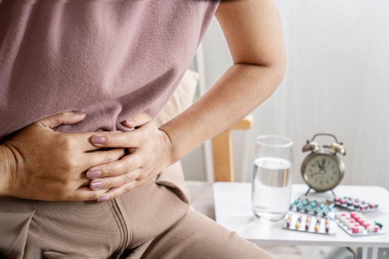 A person clutching their stomach in pain sits by a table with a glass of water, pills, and an alarm clock, wondering: what are the 3 types of IBS?.