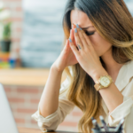 A woman sits at a desk, eyes closed and hands pressed to her face, appearing overwhelmed—perhaps searching for research verified migraine relief reviews on her laptop.