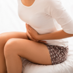A woman sitting on a bed, clutching her abdomen with both hands, appears to be in pain after waking up gassy and bloated.