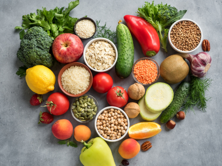 Assorted fresh fruits, vegetables, grains, and legumes are arranged in a heart shape on a gray surface, symbolizing eating habits for aging well.