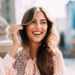 A smiling woman with long, wavy brown hair enjoys a sunny day outdoors, radiating the vitality linked to Ashwagandha With Black Pepper Benefits.