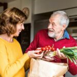 Two older adults in a kitchen, holding vegetables, discussing healthy food choices for hair growth and vitality.
