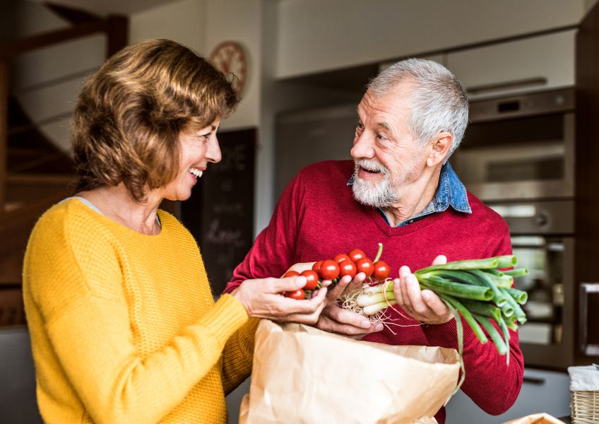 Two older adults in a kitchen, holding vegetables, discussing healthy food choices for hair growth and vitality.
