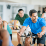 A group of people smiling and exercising together on stationary bikes in a brightly lit fitness studio, staying motivated with supplements for new year's resolutions.