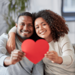 Smiling couple sitting on a couch, hugging and holding a red paper heart towards the camera, celebrating love and heart-boosting supplements.