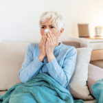 An older woman with short gray hair sits on a couch, wrapped in a blue blanket, holding a tissue to her nose—she may be considering supplements to boost immunity.
