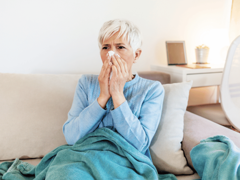 An older woman with short gray hair sits on a couch, wrapped in a blue blanket, holding a tissue to her nose—she may be considering supplements to boost immunity.