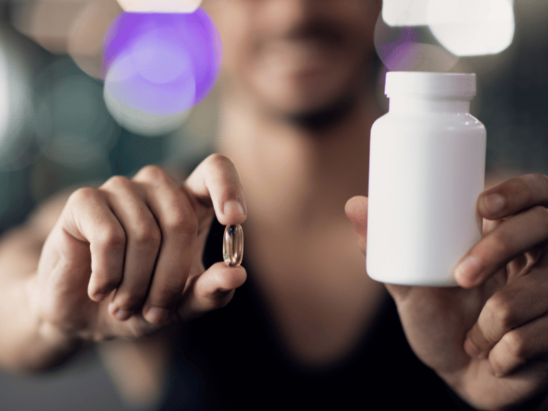 5 Surprising Supplements That Support Gut Health A person holds a white pill bottle in one hand and a single capsule—possibly supplements for gut health—in the other, with a blurred background.