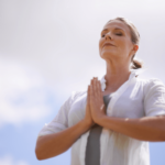 A woman stands outdoors with eyes closed and hands pressed together in a prayer position, appearing as calm and peaceful as if supported by adaptogens under a bright sky.