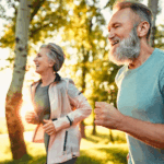 Two older adults, a man and a woman, are jogging outdoors in a sunny park, smiling and enjoying the bright, green surroundings.