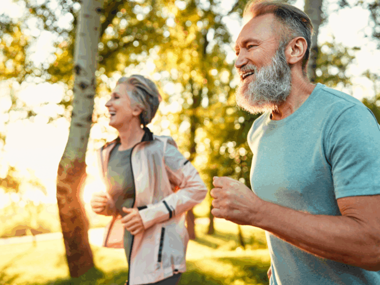 What Muscle Loss Really Costs You Two older adults, a man and a woman, are jogging outdoors in a sunny park, smiling and enjoying the bright, green surroundings.
