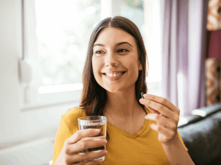 Mood Matters: The Best Supplements to Help You Feel Your Best Smiling woman in a yellow shirt holding a glass of water and a green pill, enjoying a positive mood while sitting indoors near a window.