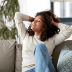 A woman sits on a couch with her hands behind her head, eyes closed, and smiling, appearing relaxed and content in a bright, cozy living room.