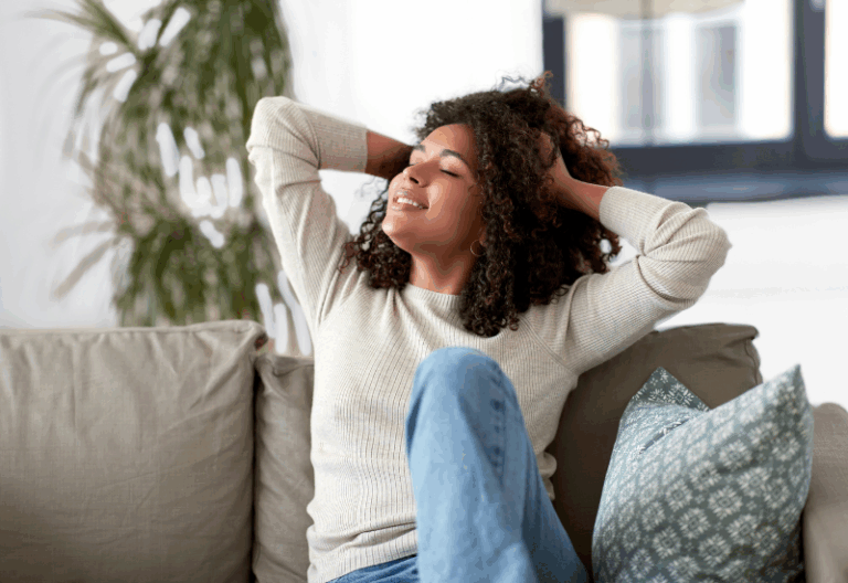 6 Simple Tips For Beating Seasonal Depression A woman sits on a couch with her hands behind her head, eyes closed, and smiling, appearing relaxed and content in a bright, cozy living room.