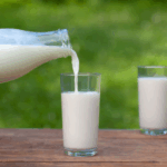 A glass bottle pours milk into a clear glass on a wooden table outdoors—making you wonder, why do I crave milk as you notice green grass and an extra glass in the background.