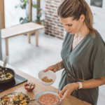 A woman in a green dress stands in a kitchen, sprinkling chickpeas onto a bowl of pink soup, with a salad and vegetables on the counter beside her.