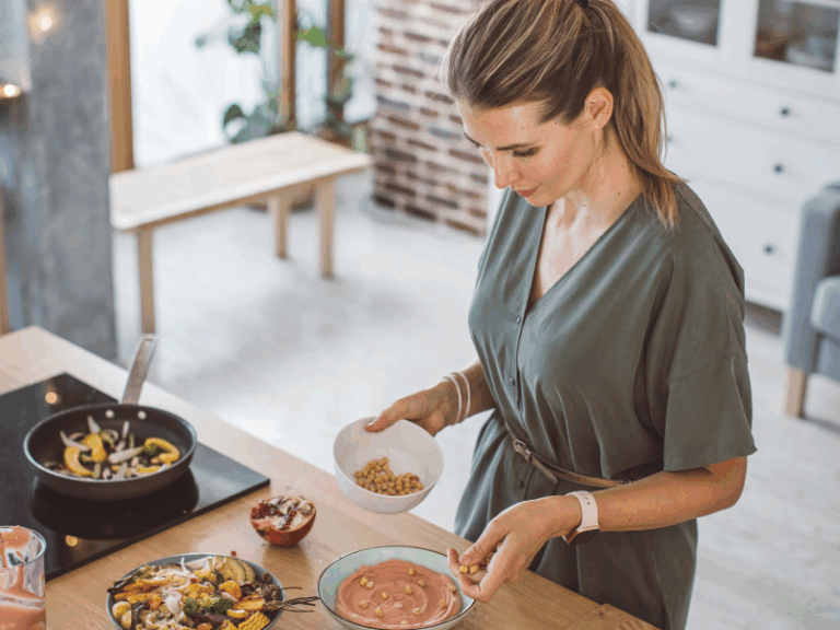 A woman in a green dress stands in a kitchen, sprinkling chickpeas onto a bowl of pink soup, with a salad and vegetables on the counter beside her.