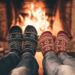 Your Holiday Check-In Guide - Two people wearing patterned winter socks relax by a fireplace during a Holiday Check-In, with mugs nearby, creating a cozy and warm atmosphere.