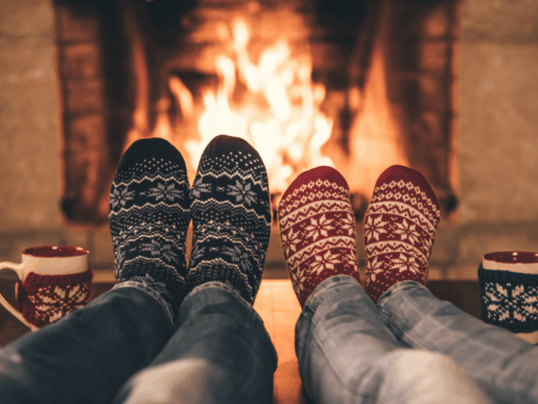 Your Holiday Check-In Guide - Two people wearing patterned winter socks relax by a fireplace during a Holiday Check-In, with mugs nearby, creating a cozy and warm atmosphere.