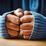 Two clenched hands resting on a wooden surface, partially covered by long blue sweater sleeves. WHAT Are Your Cold Hands Trying To Tell You?