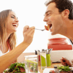 A smiling woman feeds a laughing man with a fork while they sit together at a table with food and drinks.