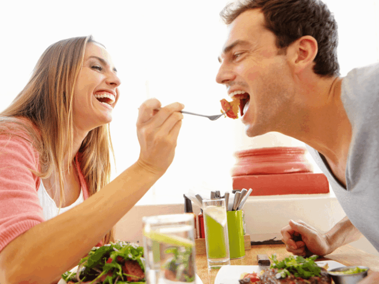 Is Eating More The Key To losing Weight? A smiling woman feeds a laughing man with a fork while they sit together at a table with food and drinks.