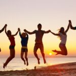 Six people are holding hands and jumping in the air on a beach at sunset, with the ocean and sun visible in the background.