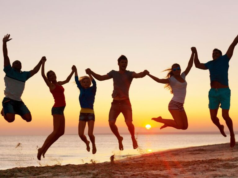 How Does Research Verified Energy Booster Work? Six people are holding hands and jumping in the air on a beach at sunset, with the ocean and sun visible in the background.