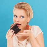 A woman with short blonde hair, battling sugar addiction, is about to eat a piece of chocolate cake, holding it close to her mouth against a blue background.