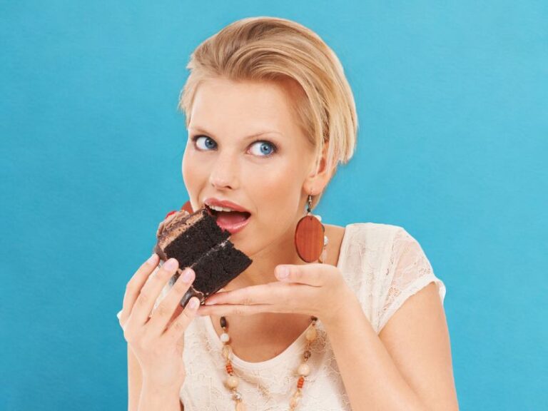 A woman with short blonde hair, battling sugar addiction, is about to eat a piece of chocolate cake, holding it close to her mouth against a blue background.