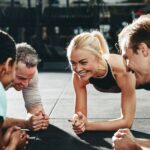 Four people are smiling and holding plank positions on a gym floor, facing each other and appearing to enjoy their workout together.
