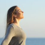 A woman with long hair stands outdoors by the sea, eyes closed, smiling peacefully and breathing deeply in the fresh air under a clear sky.