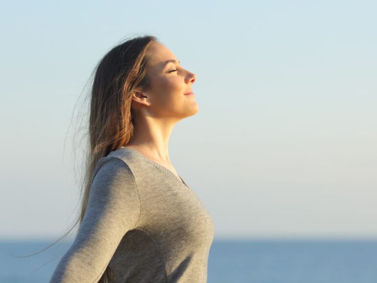 Beyond Calming: The Benefits of Breathing Exercises A woman with long hair stands outdoors by the sea, eyes closed, smiling peacefully and breathing deeply in the fresh air under a clear sky.