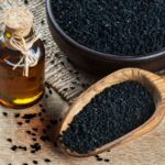 A wooden spoon and bowl filled with black seeds sit on a wooden surface next to a small glass bottle of oil with a cork and twine.