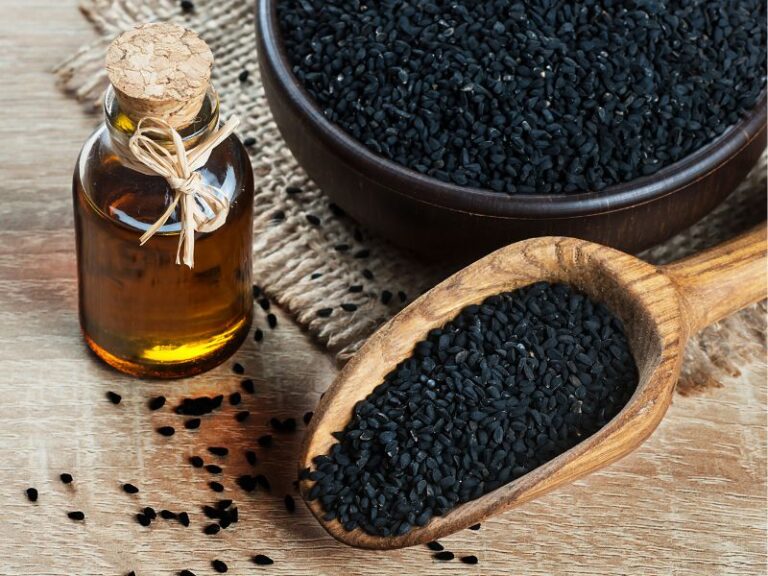 A wooden spoon and bowl filled with black seeds sit on a wooden surface next to a small glass bottle of oil with a cork and twine.