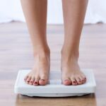 A person standing barefoot on the scale, which rests on a wooden floor, with only their feet and lower legs visible.