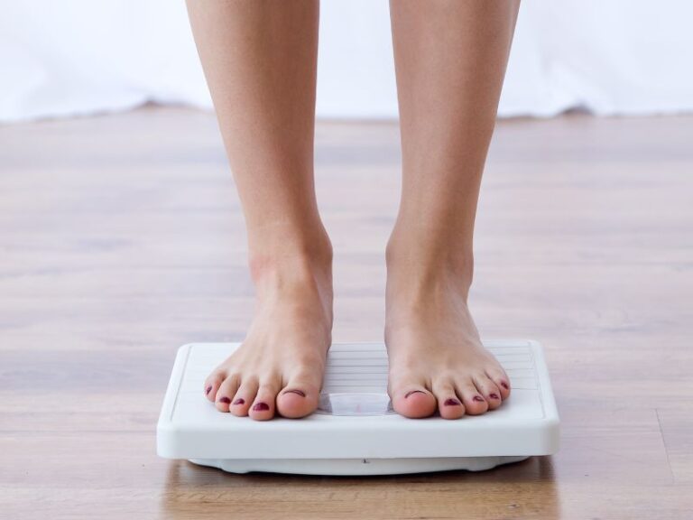 Why You Shouldn’t Trust The Scale A person standing barefoot on the scale, which rests on a wooden floor, with only their feet and lower legs visible.