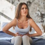 A woman sitting on a bed holds her stomach with both hands and appears to be in pain or discomfort.