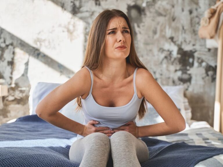 A woman sitting on a bed holds her stomach with both hands and appears to be in pain or discomfort.
