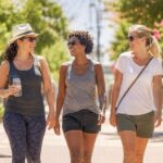 Three women wearing sunglasses and casual summer clothes walk outside on a sunny day, smiling and talking together. One holds a water bottle. Trees and flowers line the pathway.