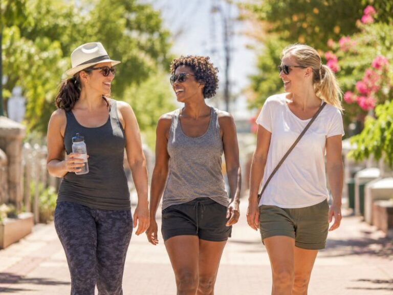 Three women wearing sunglasses and casual summer clothes walk outside on a sunny day, smiling and talking together. One holds a water bottle. Trees and flowers line the pathway.