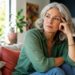 A thoughtful older woman with gray hair sits on a couch, resting her head on her hand, looking out a window. She wears a green shirt and looks pensive.