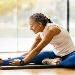 Older woman in athletic wear sits on a mat, stretching her leg and smiling in a bright room with large windows.