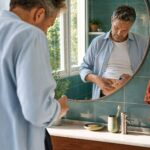 A man stands in a bathroom injecting himself in the stomach, reflected in a mirror. There are toiletries and a plant on the sink.