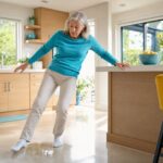 An older woman in socks slips on a wet kitchen floor, looking alarmed. She reaches toward the counter to catch herself.